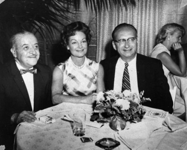 Frank Chimera, Rochelle Hudson and Robert Bloch (in eyeglasses) are seated at a table at an event related to the release of the film <i>Strait Jacket</i>. A pack of cigarettes, matches and an ashtray are on the table.