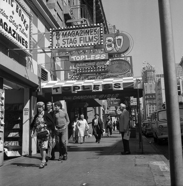 Harold Gauer standing on the sidewalk in front of a topless club, shading his eyes to look into the establishment. Signs read "Erotic Magazines Stag Films,""Dancing Girls,""Live Adult  Entertainment," and "Topless Dancing Girls."