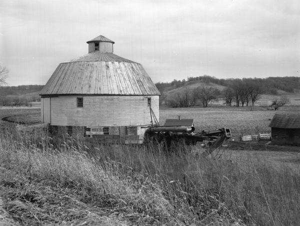View of a round barn along Highway K. Farm equipment and another farm building are on the left. Fields and hills are in the background.