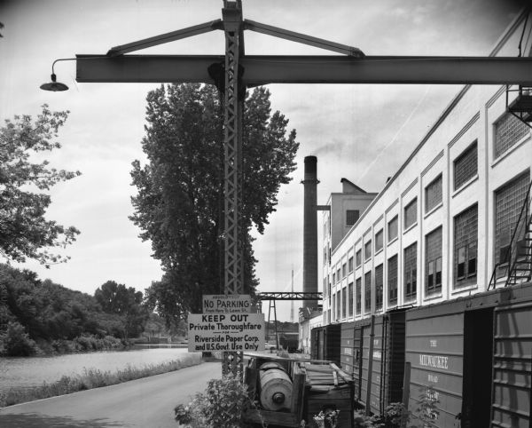 Exterior view of Riverside Paper Corporation mill. On the left is a road along a river, and on the right are railroad tracks along the mill. A sign in the foreground reads in part: "Keep Out / Private Thoroughfare." In the far background appear to be a set of locks or a dam.