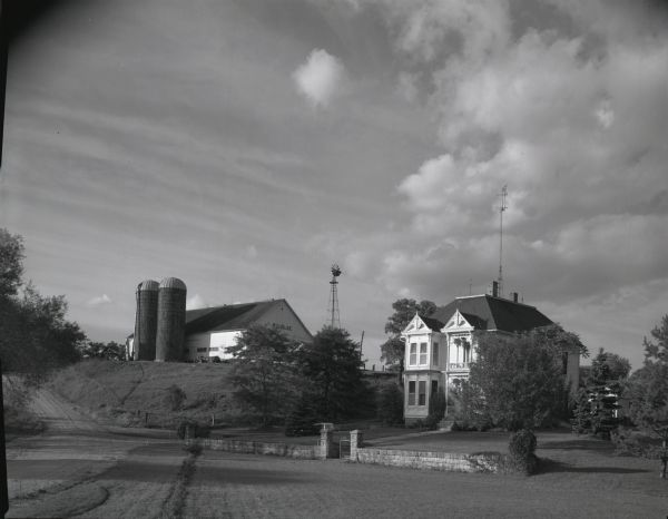 View from field of the W. Blise farm. A barn with two silos, a windmill and a large house are visible. A stone wall landscapes the area in front of the house.