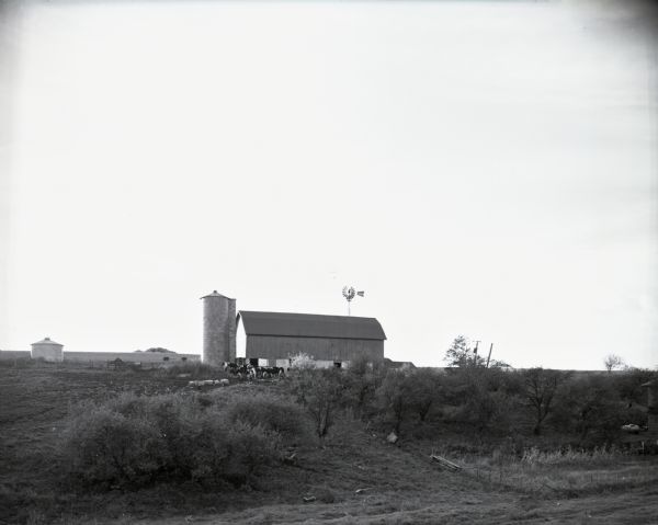 View from a distance of a dairy farm on top of a hill. A windmill, silo, barn and dairy cattle are visible.