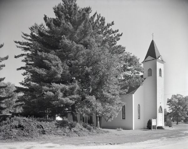 View from road of All Saint's Church behind a group of trees. The front entrance is on the right, with a signboard nearby listing times of the masses and confessions. On the right a house and outbuildings can be seen through trees.