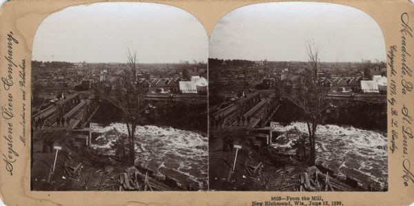 Elevated view of a town with a railroad bridge crossing over a river. Several individuals are visible crossing the bridge. Sign near bridge says: "Willow River." Caption on stereograph reads, "9665-From the Mill, New Richmond, Wis., June 12, 1899."