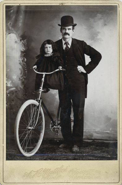 Full-length studio portrait in front of a painted backdrop of a man and a child with a large bicycle.