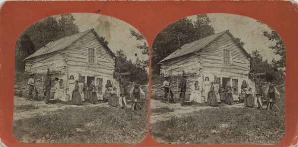 A group of men, women and children are gathered outside a log building. Two men are holding tools. Residence of Ole Nyhuus (sp?).