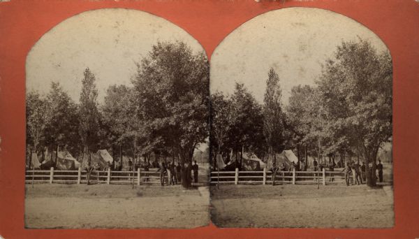 A group of men dressed in uniform gather near a fence in a shaded encampment. Several tents are visible in the background. Caption on back of stereograph reads, "Views of Camp Fair, Randall Park, Eau Claire. Time of the Strike, July 28, 1881." The strike was a mill strike  known as the "Sawdust War".