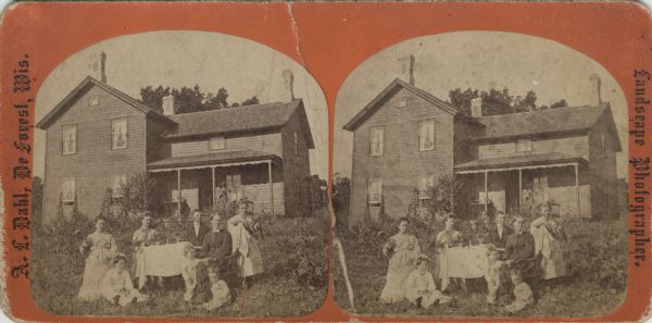 A family poses for a portrait outdoors in front of their house. The eight individuals gather around a table on their front lawn for the photograph. Caption on back of stereograph reads: "Mrs. Beret H. Stugaard and Familie[sic], Springdale, Wisconsin."