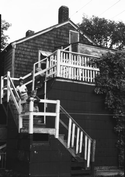 Two children stand on an outdoor stairway of wood-shingle house. Caption on back of print reads, "Back steps in 'Goosetown.' a section around Broadway and 9th Street, an old residential island surrounded by industrial plants and bare flats."