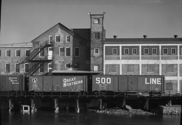 View of Gilbert Paper Company from a railroad bridge on the Fox River. Three rail cars are stopped in front of the factory.