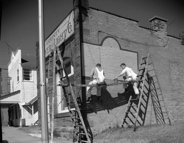 Two painters sit on an elevated, wooden plank suspended between two ladders. They are painting the side of the "County Power and Light Co" building. The building was painted to be used in a motion picture location set.