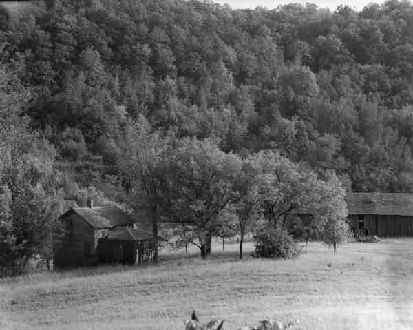 View from slope of an abandoned farm with empty farmhouse and barn in a open area surround by trees and hills.