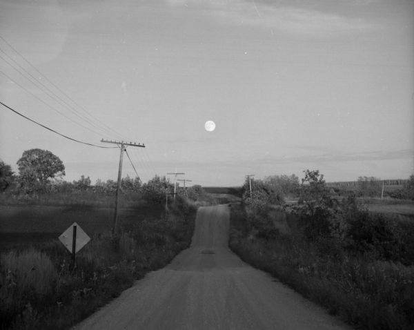 View down a winding, hilly country road. A full moon is in the background, just above the horizon line.