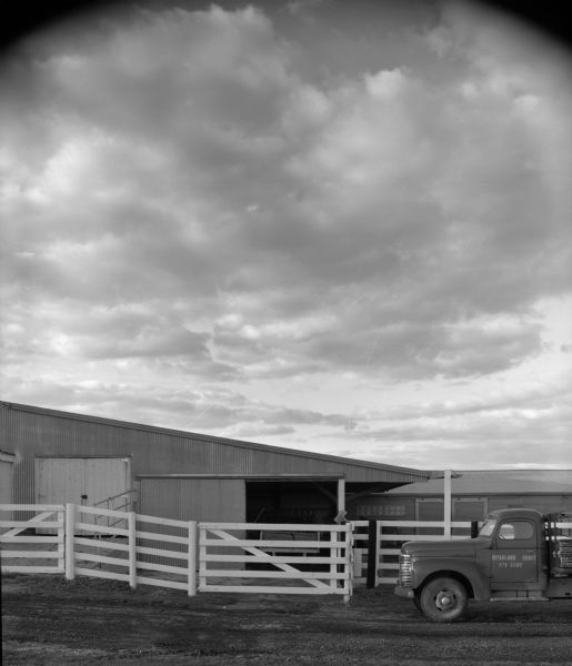 A pickup is parked outside two agricultural buildings on a dairy farm. A white fence encloses the two buildings.