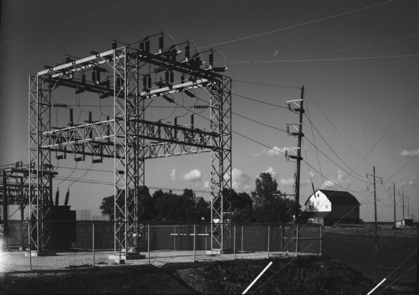 View of a rural electric substation, with a nearby farm in the background.