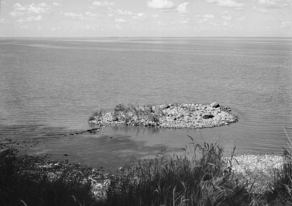 Elevated view of a small, gravelly island in Lake Michigan. Caption on photograph reads, "Green Bay shore with small gravely island. This is very likely the precise spot where Jean Nicolet came ashore to see the Indians, the first white man to set foot in Wisconsin. Exposure from the middle of the cleft in the steep bank leading down to the shore; if we accept that the Indian village was located on top of the bank or bluff at approximately this point, this cleft would be the natural passage up from the shore, or approach to the village."