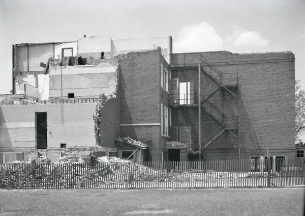 Exterior view of an old high school building, partly demolished. There is a snow fence in front of the debris.
