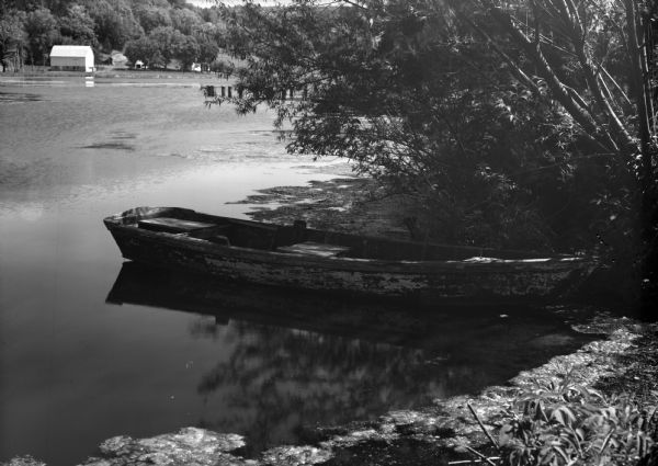 View from shoreline of an old rowboat in an artificial lake built by the Honey Creek Rod and Gun Club. There is a barn on the far shoreline.