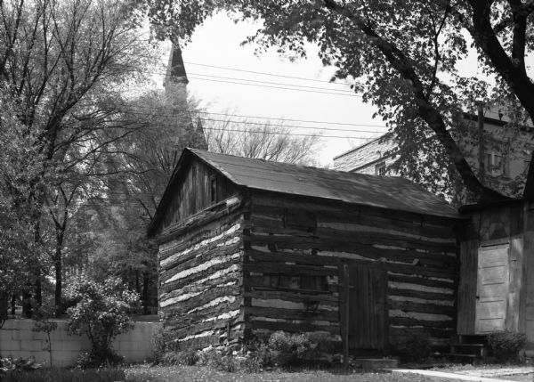 Exterior view of an early settler's log cabin. The cabin resides on private property, in downtown West Bend. The steeple of a church is in the background.