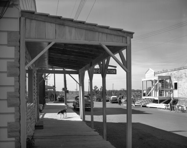 View of a downtown area reconstructed for a movie location set. Several contemporary automobiles are seen in the background. Men are working on on two different storefronts, and a dog stands on a boardwalk in the foreground.