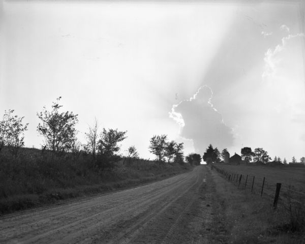 View down a country road with fences on both sides. In the distant sky, a cumulonimbus cloud is backlit by the sun. A farm with a barn and silo is on the right.