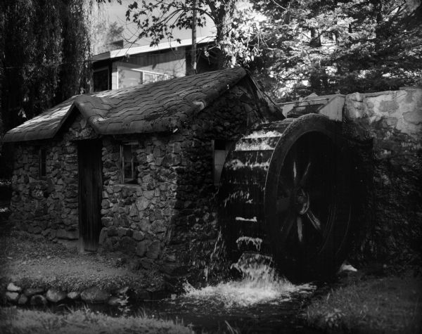 A small, stone house with a miniature water wheel. Caption on negative jacket reads, "Miniature water wheel which generates electricity to light the residence on the property of George Heubner. Mr. Heubner built the miniature mill, dam, millrace and wheel himself.