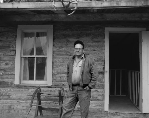 Rollo Jamison stands outside a horizontal log building. A set of deer antlers are mounted on the eave above Mr. Jamison. There is a farm implement behind him near a window.