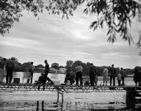 A group of men, women and children fish from a dock located on the Fox River. In the background are commercial  buildings along the shoreline.