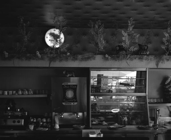Interior of Bush's Cafe, with artificial greenery, a clock, and two water pots above the lunch counter.