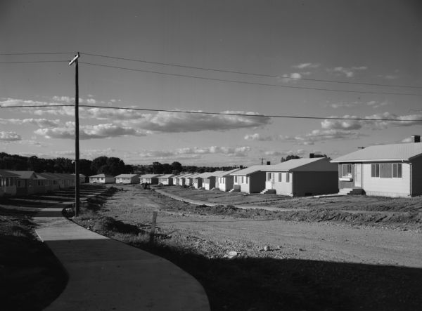 View down curving sidewalk of new housing development with a main street of dirt, and box-like, one-story homes.