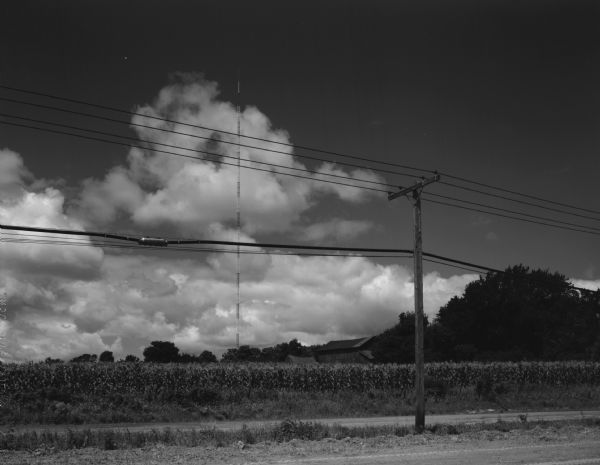 View towards a field of corn and a farm and the power lines which run along a road. A tall industrial tower rises in the background.