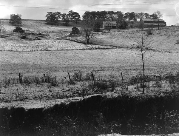 View across field of a farm on a tree-lined hillside.