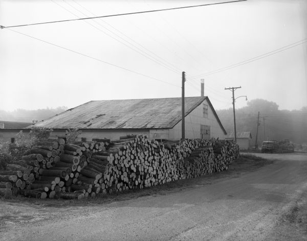 Three large rows of stacked logs are piled outside a small factory building.