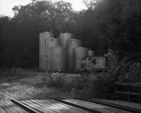 View across a railroad bridge of a group of seven oil storage tanks in a wooded area.