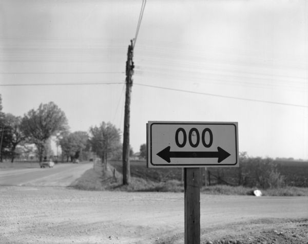 The Highway OOO sign at a road junction. Several automobiles are visible in the background along a road.