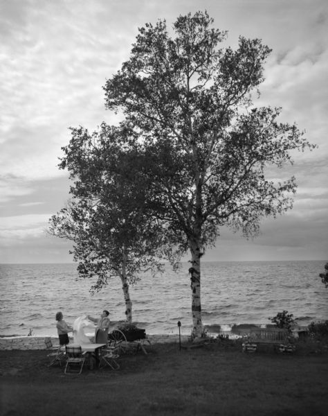 View of Lake Michigan as seen from the lakefront property owned by Mr. Lloyd Lehman. Mrs. Lehman, and a neighbor, Patti O'Connell, attempt to set a dinner table in the wind.