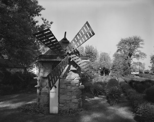 A decorative windmill in a residential garden. Large trees are in the background.