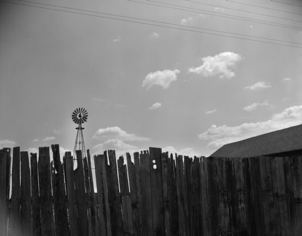 A wooden fence borders a farm. A windmill and barn rooftop are visible above and behind the fence.