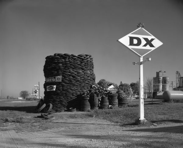 A large pile of used tires near a DX gasoline sign. In the background are several grain elevators, a house, and a Pabst Blue Ribbon beer sign.