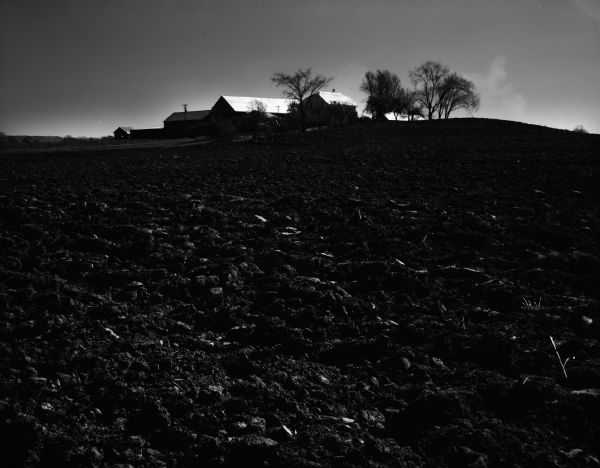 A plowed field with several farm buildings and trees in the distance.