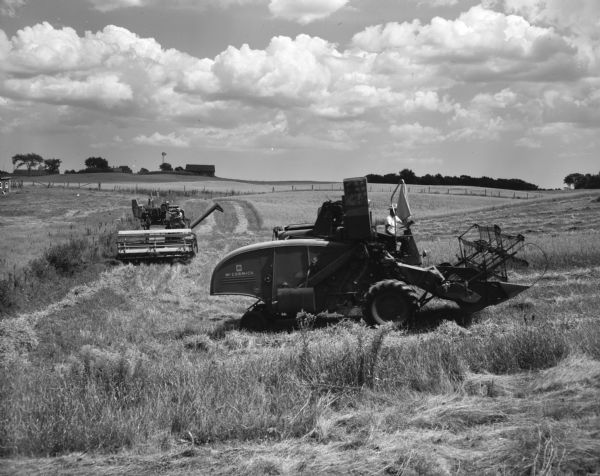 Two men on two McCormick reapers at work in a wheat field. In the distance several farm buildings are visible with large clouds in the sky.
