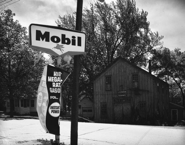 A Mobil gasoline service station sign is in the foreground. Across the street is a blacksmith shop. On the left is a residential home next to the blacksmith shop, surrounded by trees.