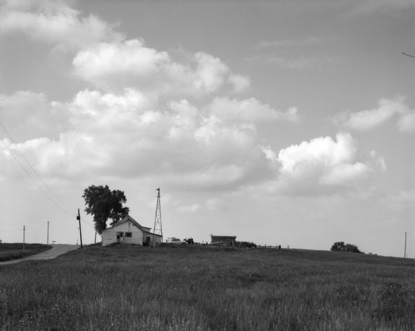 Landscape view from field of a rolling countryside. A small cheese factory resides on a slope, with a tree, a windmill, two cars and a tractor near an outbuilding.