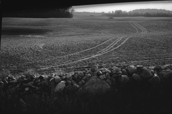 View over rock wall of a plowed field with tractor tracks in the dirt.