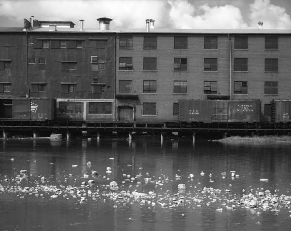 View across water of a railroad bridge along the side of a strip of industrial buildings. Railroad cars are stopped in front of the buildings.