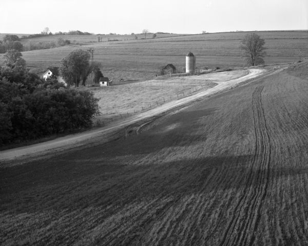 Elevated view over plowed field and dirt road toward a farm with a farmhouse, barn, silo and other outbuildings residing in a sloping valley.