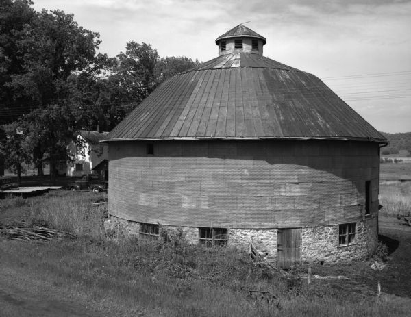 Exterior view of a farm with a round barn. A truck and farmhouse is behind and to the left in a grove of trees.