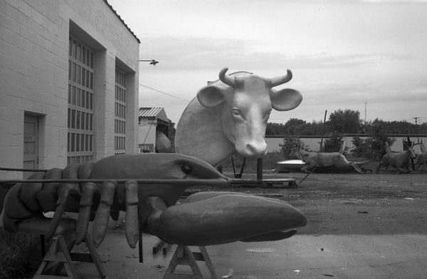 Sculptured Advertising Company (F.A.S.T. Corporation of Sparta is an outgrowth of Sculptured Advertising) with unfinished prop displays, perhaps made of fiberglass, outdoors near the garage doors of a building. In the foreground are a large cow head and lobster. Several prop deer are in the background.
