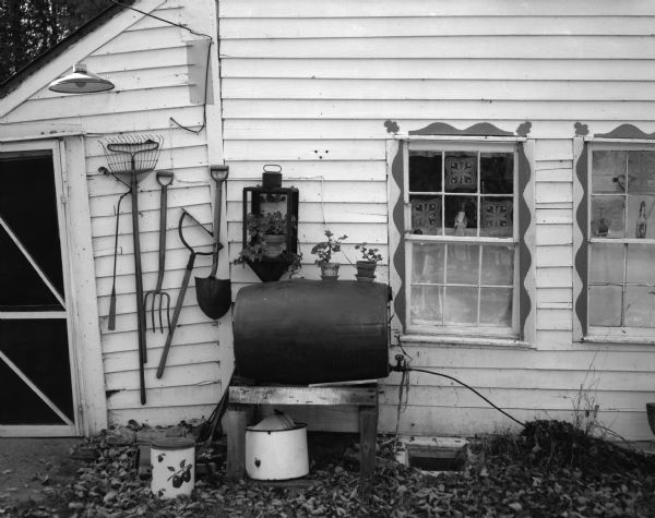 View of a side wall and rear entrance of a small country home owned by Mrs. Kay Curtis. A collection of yard tools hang from the siding near a screen door, and potted plants sit on top of a water barrel near two windows.