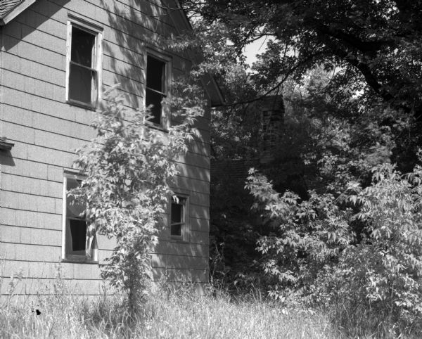 Side view of a deserted farmhouse, surrounded by trees. In the background, the older, rear wing of the house is visible.